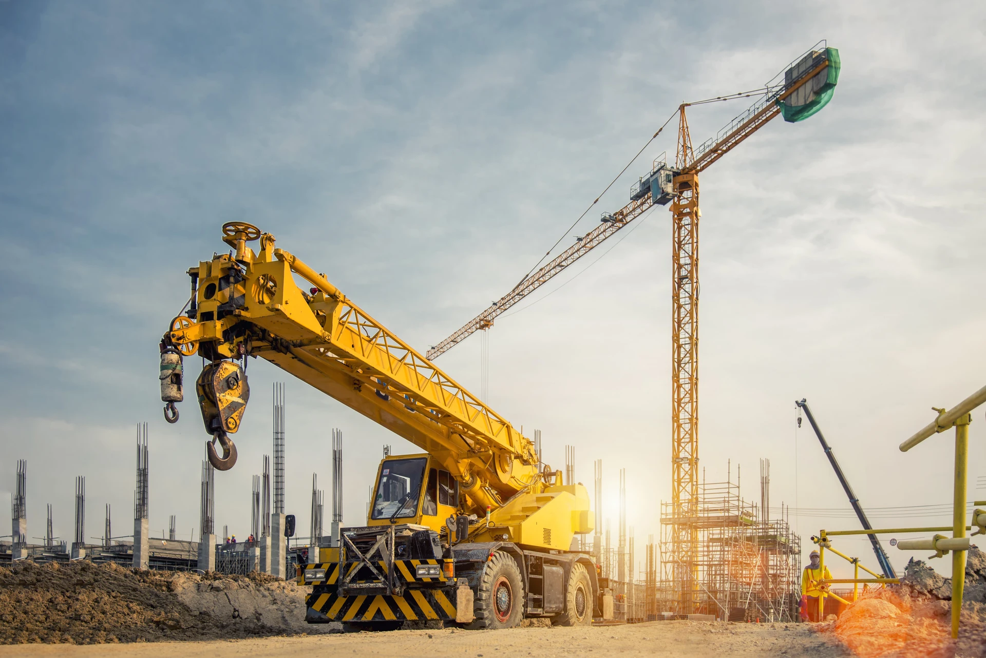 Chantier de construction avec grue mobile jaune au premier plan, grue à tour en arrière-plan, structures en béton armé avec ferraillage apparent