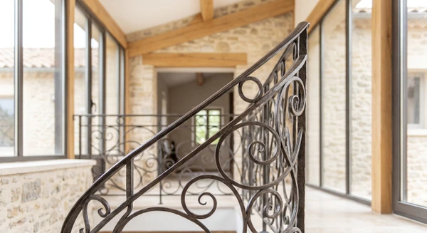 Détail photographique d'une création en fer forgé ouvragé : escalier hélicoïdal avec rampe en fer forgé aux motifs arabesques et volutes forgées à la main, structure tubulaire noire ou patine rouille, marches en métal ou bois, plan de détail rapproché montrant la qualité artisanale des finitions et des motifs décorés. Environnement intérieur lumineux de type escalier contemporain ou vérandas. Photographie professionnelle en haute définition soulignant la qualité du travail artisanal et des détails décoratifs.