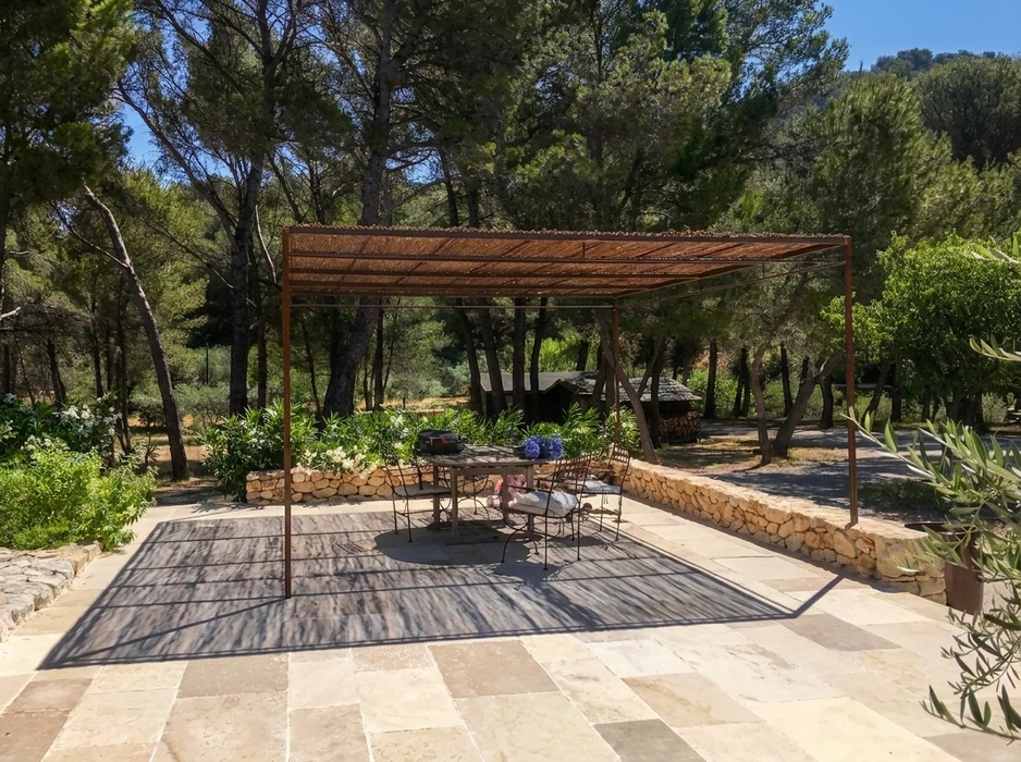 Terrasse extérieure en pierre calcaire avec pergola métallique à toit en cannisse, abritant une table ronde et chaises en fer forgé