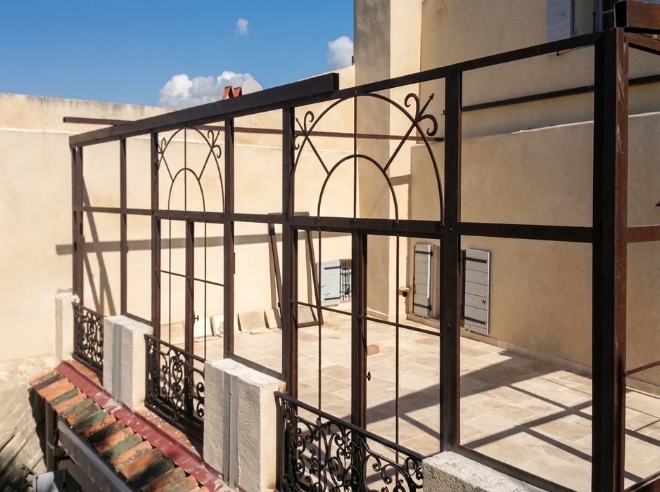 Terrasse extérieure d'un bâtiment méditerranéen avec pergola en fer forgé brun-rouille aux motifs décoratifs en arabesques et volutes