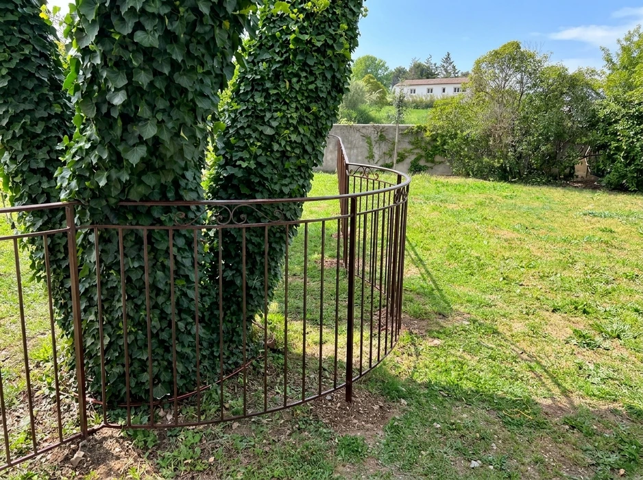 Jardin extérieur verdoyant avec une clôture en fer forgé marron foncé à barreaux verticaux, courbée en arc de cercle autour d'un pilier recouvert de lierre