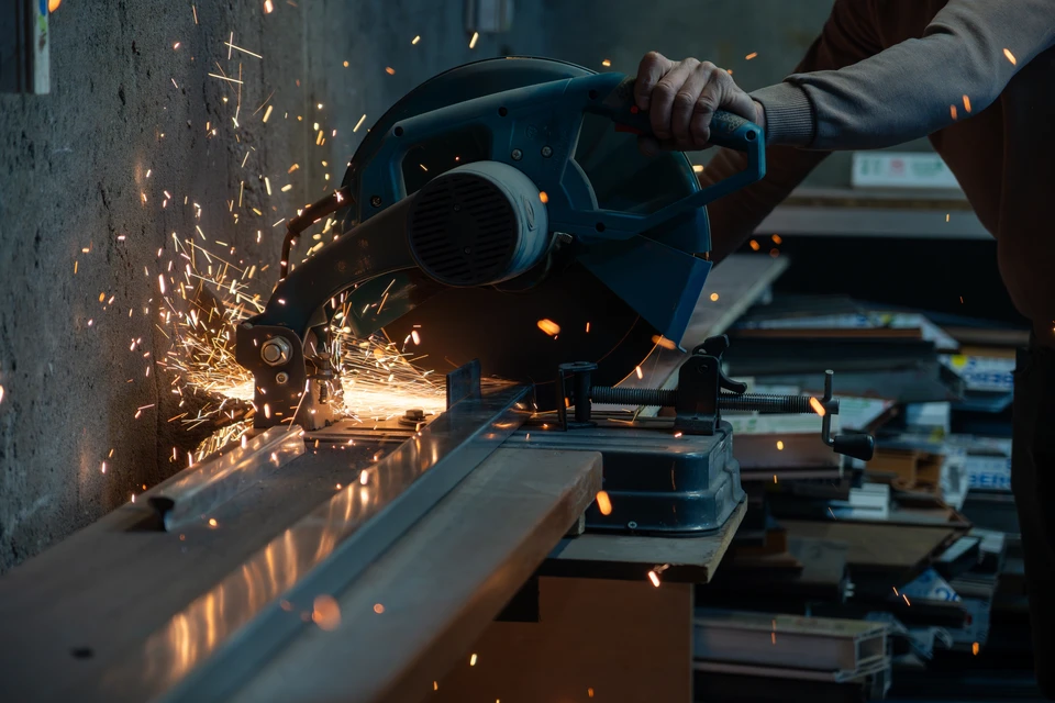 Photo réelle d'un artisan ferronnier en tenue de travail professionnelle (vêtements de sécurité appropriés) en train de travailler sur une réalisation métallique dans l'atelier ou sur un chantier du Groupe Bassé. L'artisan manipule des éléments métalliques (portail, pergola, garde-corps) avec des outils professionnels. On distingue clairement l'expertise manuelle et l'environnement de travail artisanal. Lumière naturelle, ambiance de professionnel au travail concentré sur sa tâche. Format horizontal, pas de texte ni logo visible.
